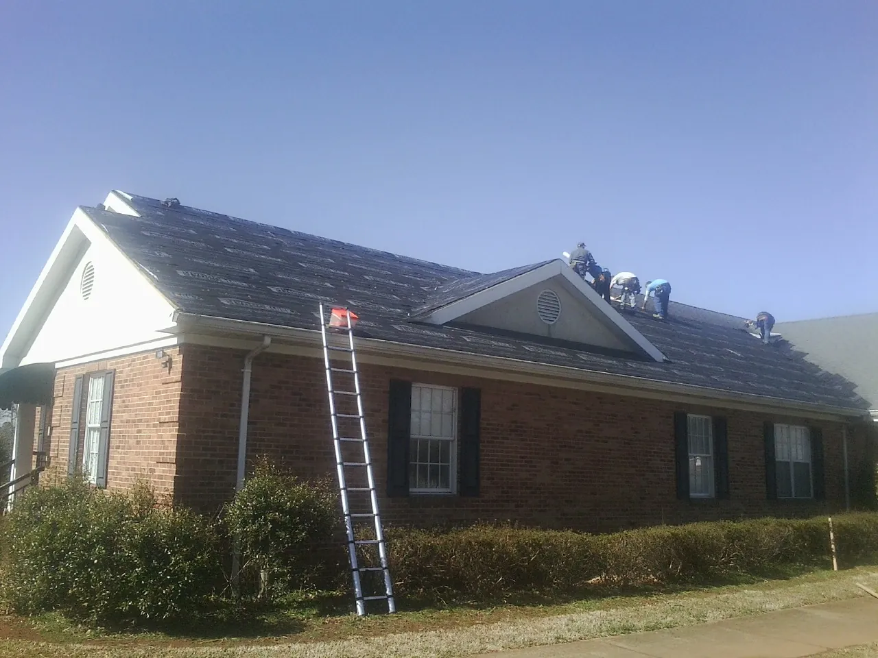 Crew tearing shingles off a brick ranch roof with a ladder set — Emergency Roof Repair work by our crew in Matthews
